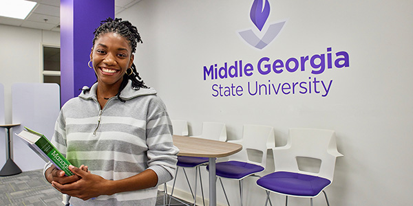 Image of Kanaiya Cornelius posing smiling for a photo holding a green textbook in front of an MGA logo in a white-walled room.
